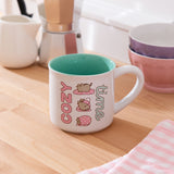 Ceramic Pusheen Mug sits on a brown desktop. The mug features Pusheen designs on each side of the mug.