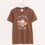 Pusheen Iced Coffee Tee hangs on a light pink hanger on a light background. The medium brown tee shirt has a graphic of Pusheen the Cat atop her favorite iced coffee beverages and some donuts.