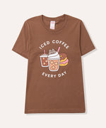 Coffee t-short lies on a flat surface. The brown tee has a large graphic in the center front of the t-shirt feauring Pusheen the Cat, two iced coffees, two iced donuts, and the phrase "Iced Coffee Every Day."
