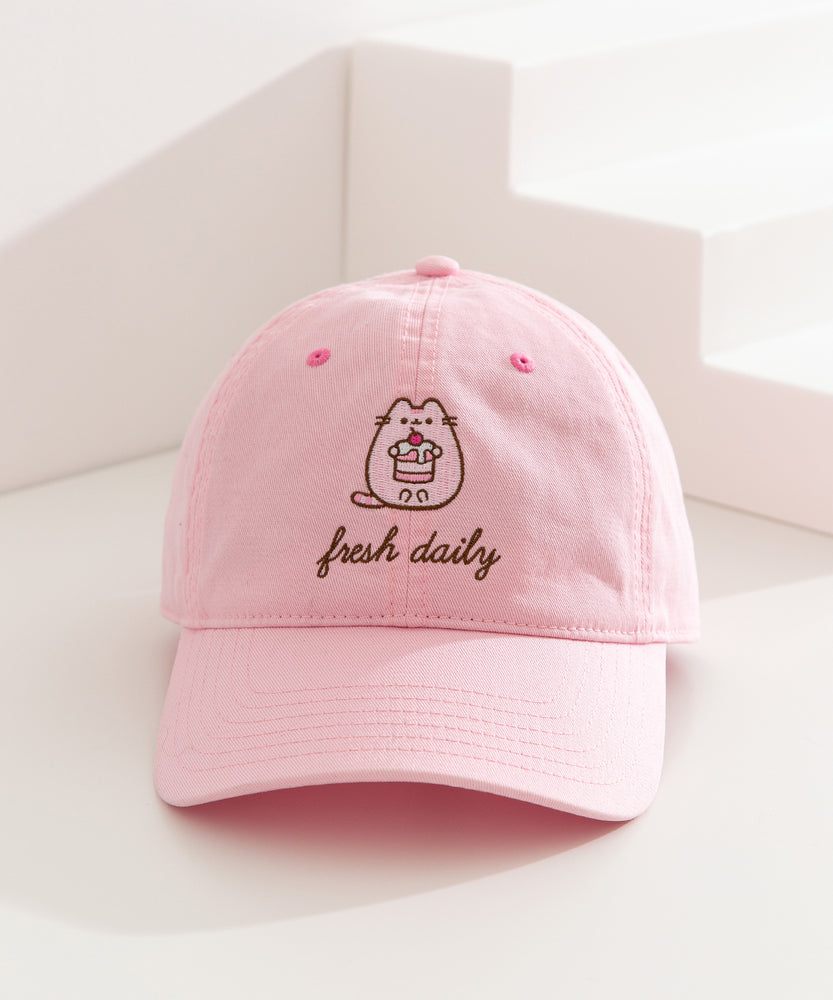 Pusheen Sweets Cap on a white surface. The light pink cap features Pusheen the Cat in a pink hue while outlined by a dark brown color. The phrase "fresh daily" sits below the cat embroidery. 