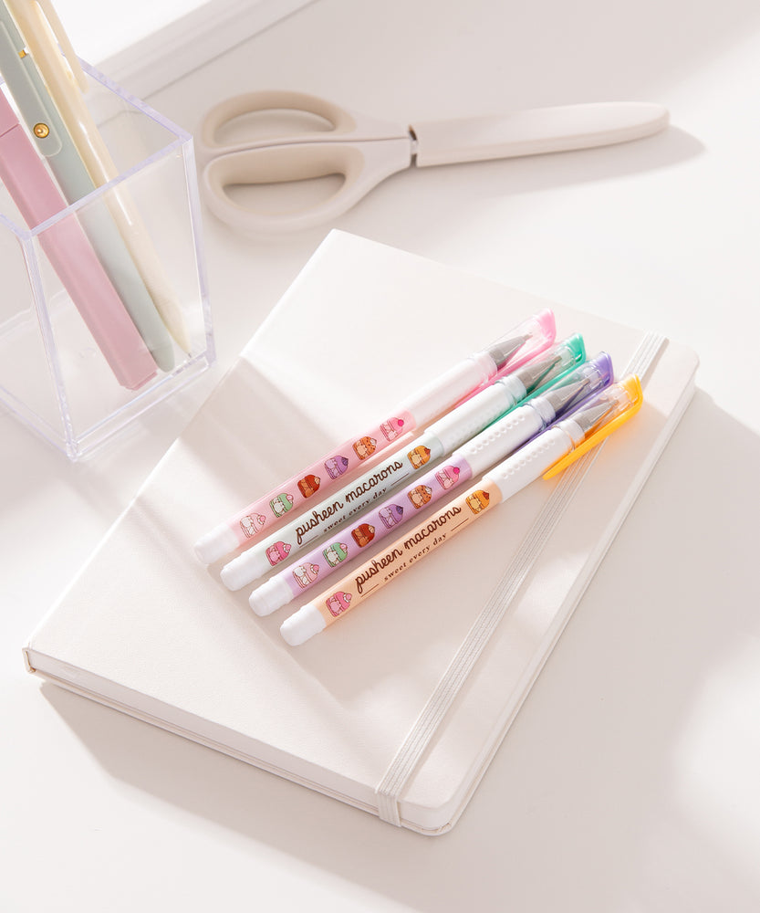 Pusheen Sweets Gel Pen set lies on a white notebook. The four pens have pink, green, purple, and orange barrels with pusheen macaron graphic patterns.