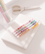 Pusheen Sweets Gel Pen set lies on a white notebook. The four pens have pink, green, purple, and orange barrels with pusheen macaron graphic patterns.
