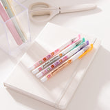 Pusheen Sweets Gel Pen set lies on a white notebook. The four pens have pink, green, purple, and orange barrels with pusheen macaron graphic patterns.