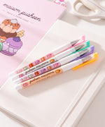 Pink, green, purple, and orange barreled pens lie on a white notebook. The four pens have macaron and Pusheen graphics printed down the barrels of each pen.