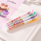 Pink, green, purple, and orange barreled pens lie on a white notebook. The four pens have macaron and Pusheen graphics printed down the barrels of each pen.