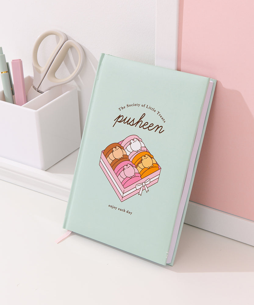 Pusheen Notebook rests on a wall to show the size of the notebook and the amount of lined pages included the luxury notebook.