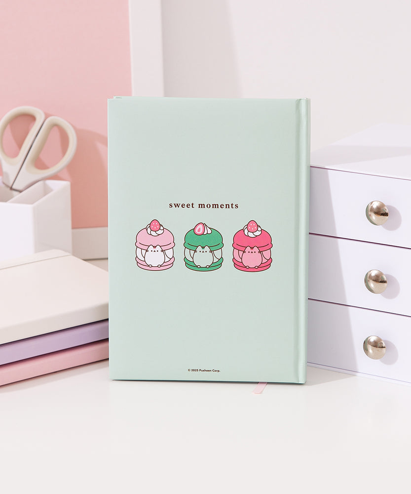 Back cover of the mint green Pusheen notebook. On the center of the back are three graphics of Pusheen the Cat transformed in macarons. On the left is a pink and white vanilla macaron, in the middle is a two-toned green matcha macaron, and on the right is a two-toned pink strawberry macaron. 