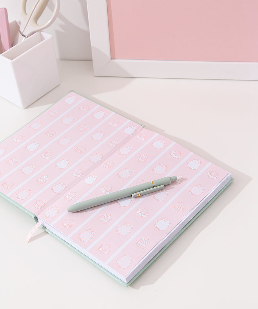 Interior view of the notebook. The inside cover features a pink striped print with macarons, cakes, whipped creams topped with strawberries and two colored Pusheen the Cats in a sitting pose.