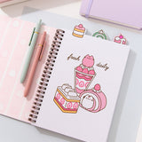 The cover of the notebook opened to the first page. The interior cover features a pink striped pattern of Pusheen with cakes, macarons, and whipped cream. The other page has a tab of a mini strawberry cake. The graphic on the right page features Pusheen transformed into a multi-pink strawberry parfait, swiss roll, and rectangular tiered cake.
