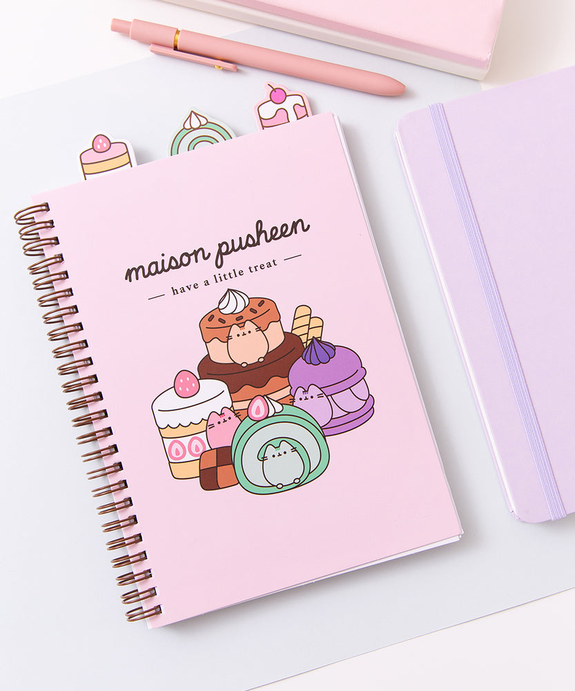 Pusheen Sweets Notebook lies on a desktop. The light pink spiral notebook has graphic of various colorful desserts including a green matcha swiss roll and multi-brown chocolate cake.