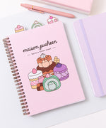 Pusheen Sweets Notebook lies on a desktop. The light pink spiral notebook has graphic of various colorful desserts including a green matcha swiss roll and multi-brown chocolate cake.