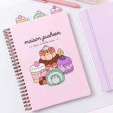 Pusheen Sweets Notebook lies on a desktop. The light pink spiral notebook has graphic of various colorful desserts including a green matcha swiss roll and multi-brown chocolate cake.