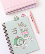The open notebook shows a green page with a large graphic of a matcha flavored parfait and swiss roll. The green desserts are accompanied by the phrase "sweet every day." Above the notebook are two tabs featuring a matcha flavored swiss roll and a cherry mini cake.