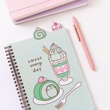 The open notebook shows a green page with a large graphic of a matcha flavored parfait and swiss roll. The green desserts are accompanied by the phrase "sweet every day." Above the notebook are two tabs featuring a matcha flavored swiss roll and a cherry mini cake.