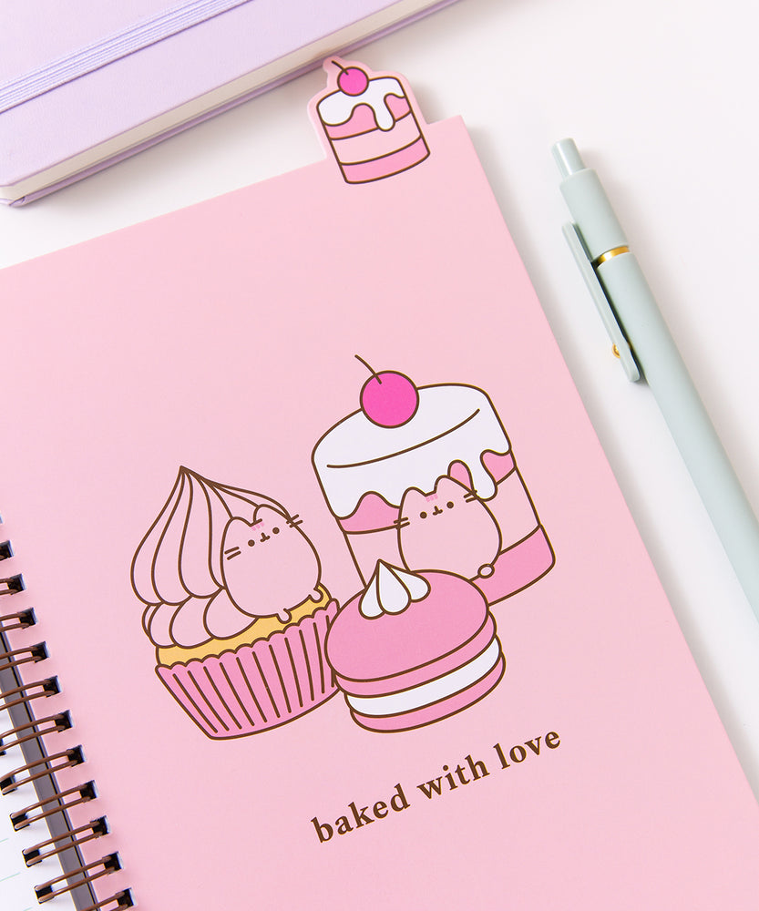 The open notebook shows a pink page with a large graphic of a cherry flavored cupcakes, cakes, and macaron The pink desserts are accompanied by the phrase "baked with love." Above the notebook is a tab featuring a cherry flavored tiered mini cake.