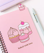 The open notebook shows a pink page with a large graphic of a cherry flavored cupcakes, cakes, and macaron The pink desserts are accompanied by the phrase "baked with love." Above the notebook is a tab featuring a cherry flavored tiered mini cake.