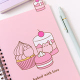 The open notebook shows a pink page with a large graphic of a cherry flavored cupcakes, cakes, and macaron The pink desserts are accompanied by the phrase "baked with love." Above the notebook is a tab featuring a cherry flavored tiered mini cake.