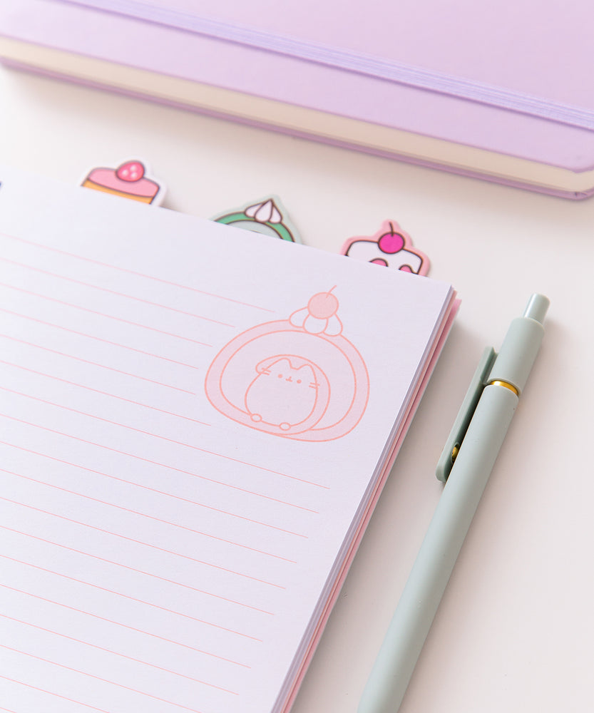 Notebook opened to a page with writing lines and a graphic of Pusheen the Cat transformed into a pink-hued cherry swiss roll at the top right of the page. The notebooks top tabs can be seen in this view.