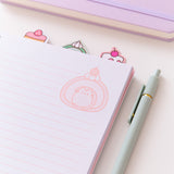 Notebook opened to a page with writing lines and a graphic of Pusheen the Cat transformed into a pink-hued cherry swiss roll at the top right of the page. The notebooks top tabs can be seen in this view.