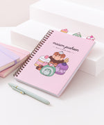 A light pink spiral bound notebook rests on  a white stair display. The large graphic on the front cover shows Pusheen as four different desserts including cakes, a macaron, and a swiss roll.
