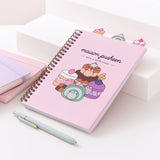 A light pink spiral bound notebook rests on  a white stair display. The large graphic on the front cover shows Pusheen as four different desserts including cakes, a macaron, and a swiss roll.