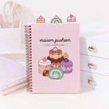 A light pink spiral bound notebook features a graphic of Pusheen as various dessert cakes and pastries. Pusheen has transformed into a large chocolate cake, mini strawberry cake, lavender macaron, and matcha-flavored swiss roll. The phrase printed above the dessert graphic is "maison pusheen, have a little treat"