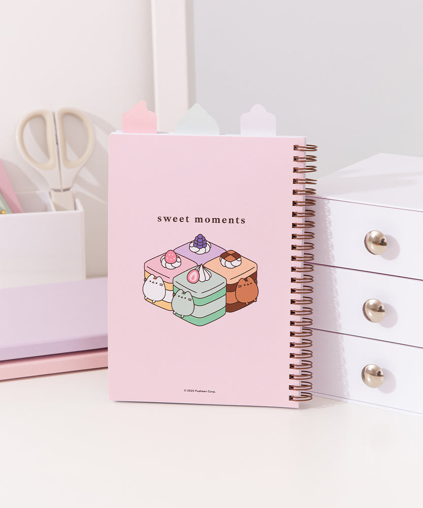 Back cover of the Pusheen Sweets Notebook. The top tabs can be seen in plain white. The back graphic shows Pusheen transformed into flavored and colorful mini cakes with the phrase "sweet moments" above. 