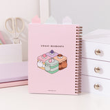 Back cover of the Pusheen Sweets Notebook. The top tabs can be seen in plain white. The back graphic shows Pusheen transformed into flavored and colorful mini cakes with the phrase "sweet moments" above. 