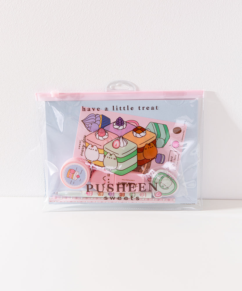 Front view of the enclosed Pusheen Sweets Stationery Set. The items in the set are encased in a clear and pink plastic resealable pouch with a graphic of Pusheen the Cat as various cakes including vanilla, lavender, chocolate, and matcha flavors.