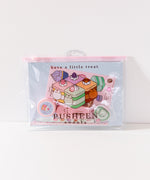 Front view of the enclosed Pusheen Sweets Stationery Set. The items in the set are encased in a clear and pink plastic resealable pouch with a graphic of Pusheen the Cat as various cakes including vanilla, lavender, chocolate, and matcha flavors.