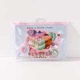 Front view of the enclosed Pusheen Sweets Stationery Set. The items in the set are encased in a clear and pink plastic resealable pouch with a graphic of Pusheen the Cat as various cakes including vanilla, lavender, chocolate, and matcha flavors.