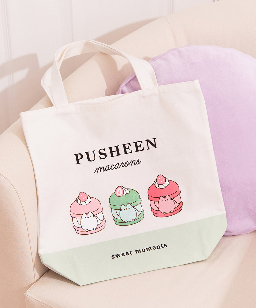 Pusheen Sweets Tote Bag lies on white chair. The white canvas tote bag features a grey Pusheen the Cat inside three sweet dessert macarons. Above the macaron graphic is the phrase “Pusheen Macarons”  and below the graphic is the phrase "sweet moments"