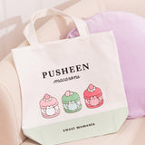Pusheen Sweets Tote Bag lies on white chair. The white canvas tote bag features a grey Pusheen the Cat inside three sweet dessert macarons. Above the macaron graphic is the phrase “Pusheen Macarons”  and below the graphic is the phrase "sweet moments"