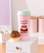 Sweets Pusheen Travel Mug on a desktop with a chocolate cupcake next to the pink Pusheen cup.