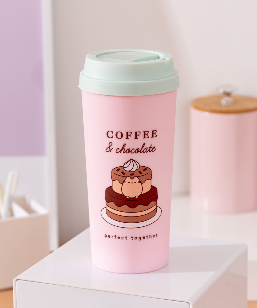 Sweets themed Travel mug sits on a desk top. The pink container has large graphics of Pusheen with a chocolate cake. The travel mug comes with a light mint green lid.