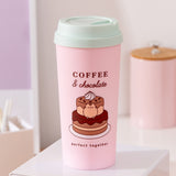 Sweets themed Travel mug sits on a desk top. The pink container has large graphics of Pusheen with a chocolate cake. The travel mug comes with a light mint green lid.