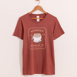 Pusheen Tea Tee hangs on a light brown hanger on a light background. The red tee shirt has a graphic of Pusheen the Cat inside a tea cup surrounded by various tea phrases and a tea bag shaped border.