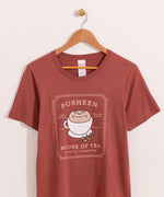 Rustic red t-shirt with a large graphic on the front center. The graphic shows Pusheen the Cat nestled in a tea cup on a saucer with a leaf sprig next to the cup.