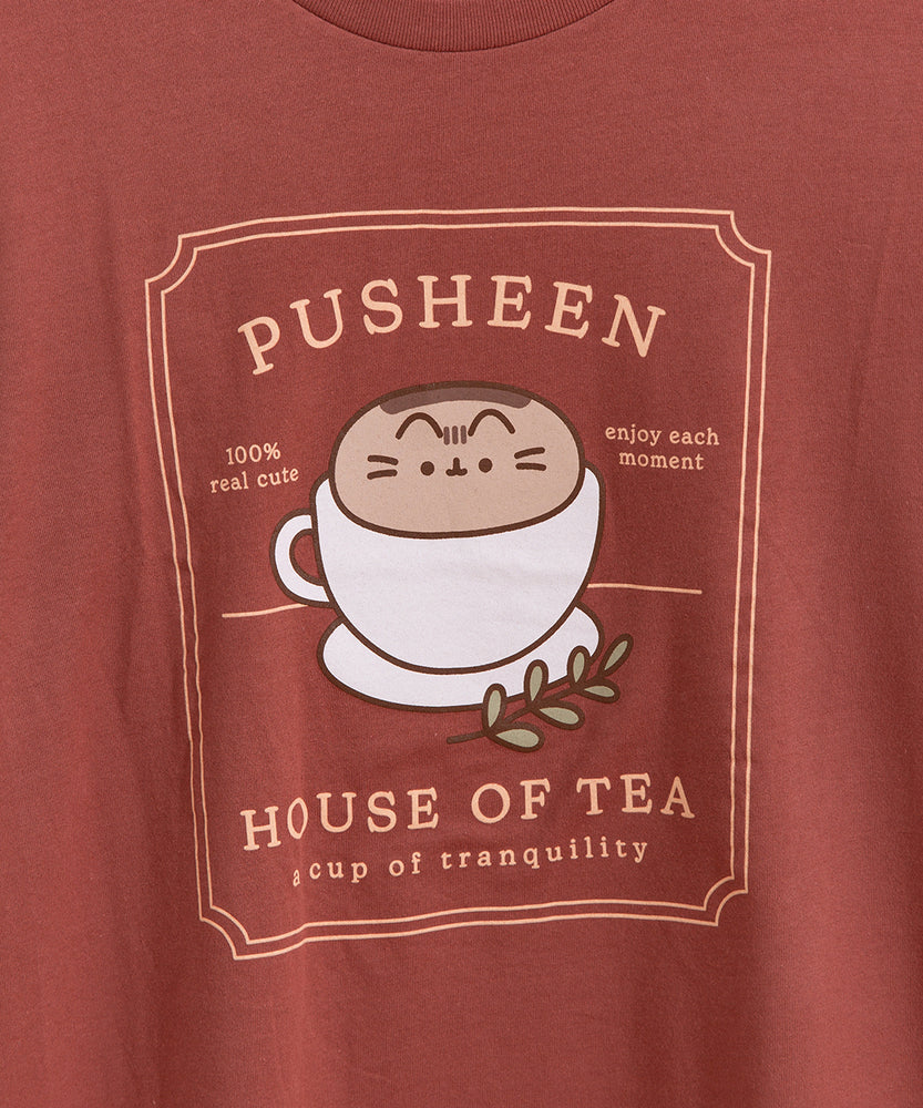 Red brown graphic tee lies on a white tabletop. The tee has a large graphic in the center of the tee featuring Pusheen the Cat in a tea cup.