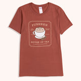 Tea t-short lies on a flat surface. The red tee has a large graphic in the center front of the t-shirt feauring Pusheen the Cat nestled inside a tea cup giving her body an extra round appearance. 