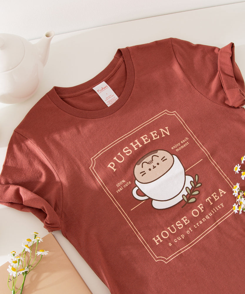 Red brown graphic tee lies on a white tabletop. The tee has a large graphic in the center of the tee featuring Pusheen the Cat in a tea cup.