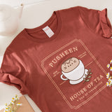 Red brown graphic tee lies on a white tabletop. The tee has a large graphic in the center of the tee featuring Pusheen the Cat in a tea cup.