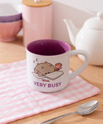 Mug with printed Pusheen graphic on each side. The white mug is placed in front of a kettle and spoon.