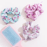 Pusheen 3-Pack Hair Scrunchies lies on a white desktop. The trio includes light pink, mint green, and white scrunchies. Each scrunchie has a different all over print of Pusheen with various icons including hearts, stars, and more. 