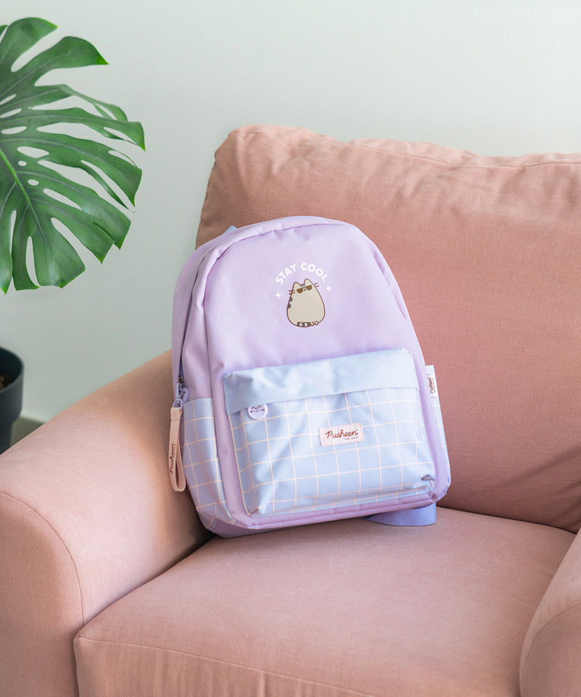 Purple backpack lies on a pink chair to show the size of the backpack. The backpack is accented with light purple details including the handle loop, brand patch, front pouch cover, zipper, and arm adjustment straps. The front graphic on the backpack is Pusheen the Cat wearing sunglasses with the phrase "Stay Cool."