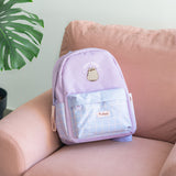 Purple backpack lies on a pink chair to show the size of the backpack. The backpack is accented with light purple details including the handle loop, brand patch, front pouch cover, zipper, and arm adjustment straps. The front graphic on the backpack is Pusheen the Cat wearing sunglasses with the phrase "Stay Cool."