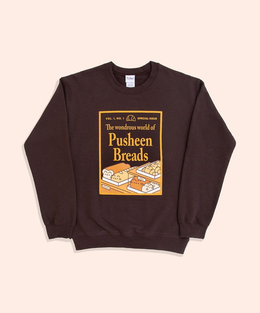 A chocolate brown sweatshirt with the sleeves folded lies against a light-yellow background. The unisex sweatshirt has a screen-printed graphic on the center of the chest. The graphic depicts a magazine cover showcasing “the wondrous world of Pusheen Breads.” This is a special, first edition cover featuring Loaf Pusheen, Baguette Pusheen, Croissant Pusheen, Loaf Pusheen, and Sesame Bagel Pusheen in golden shades of baked goodness. 