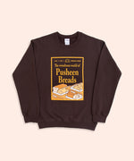 A chocolate brown sweatshirt with the sleeves folded lies against a light-yellow background. The unisex sweatshirt has a screen-printed graphic on the center of the chest. The graphic depicts a magazine cover showcasing “the wondrous world of Pusheen Breads.” This is a special, first edition cover featuring Loaf Pusheen, Baguette Pusheen, Croissant Pusheen, Loaf Pusheen, and Sesame Bagel Pusheen in golden shades of baked goodness. 