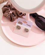 Cat-shaped earrings on a card with a scrunchie and sunglasses on a pink surface