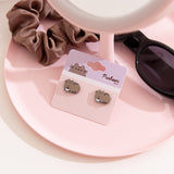 Cat-shaped earrings on a card with a scrunchie and sunglasses on a pink surface